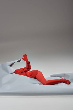 Kırmızılı, eldivenli ve taytlı bir model gri arka planda yırtık bir kağıdın yanında yatıyor.