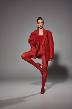 Kırmızı elbiseli şık bir kadın kameraya bakarken gri arka planda bacaklarını kaldırmış poz veriyor.