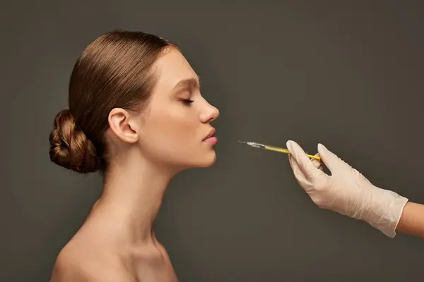 stock image esthetician in medical glove holding syringe near young woman on grey background, side view
