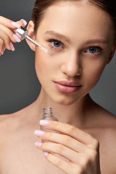 young woman with glowy skin holding cropped and bottle with serum on grey background, hydration