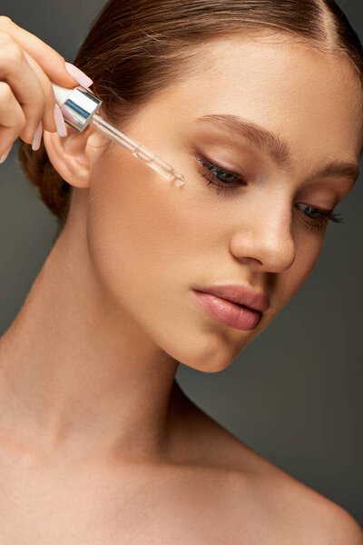 young woman with glowy skin holding dropper with serum near face on grey background, hydration
