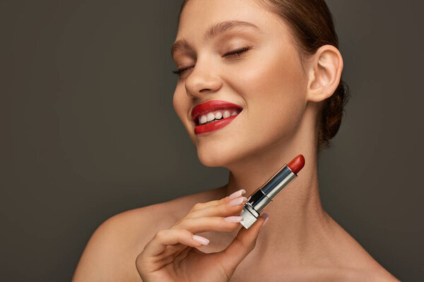 happy young woman with red lips holding lipstick and smiling on grey background, holiday makeup