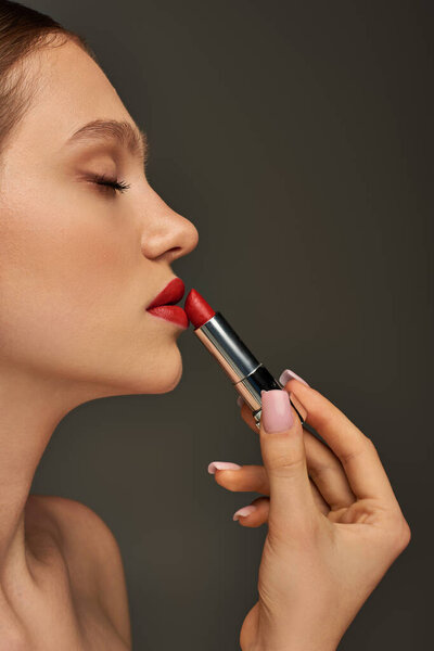 side view of beautiful young woman applying red lipstick on grey background, beauty and makeup