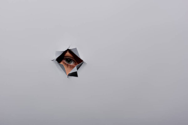 частичный снимок молодой женщины с голубым глазом, смотрящей в камеру через отверстие на сером фоне