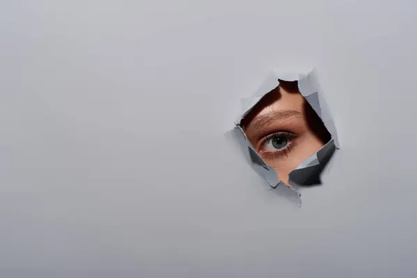 stock image cropped shot of young woman with blue eye looking at camera through hole in ripped grey background