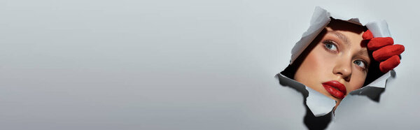 знамя молодой женщины с голубыми глазами и красной помадой, подглядывающей через отверстие в рваной серой бумаге