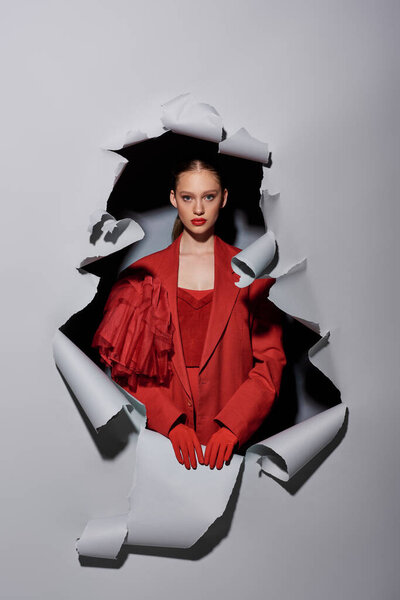 стильная молодая женщина с смелым макияжем позирует в красных перчатках возле отверстия на рваном сером фоне