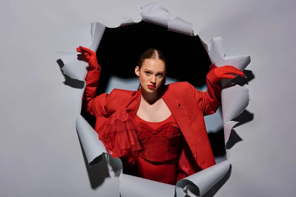 powerful young woman in red attire with bold makeup breaking through grey background with hole