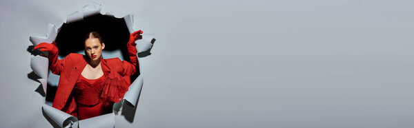 модная молодая женщина в красной одежде с смелым макияжем прорываясь через серый фон с отверстием, баннер