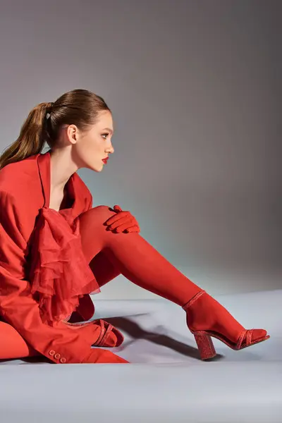 side view of stylish young model in red outfit with tights and high heels sitting on grey background