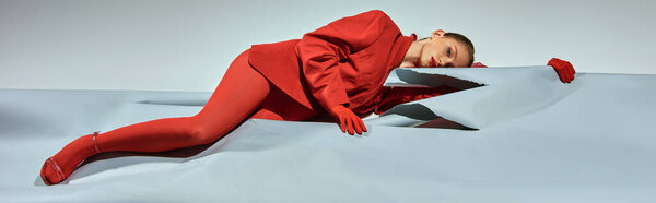 молодая женщина в красной одежде с перчатками и колготками, лежащая рядом с разорванной бумагой на сером фоне, баннер