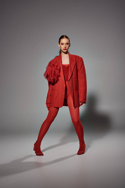 стильная женщина в красном наряде смотрит в камеру, позируя и глядя на камеру на сером фоне