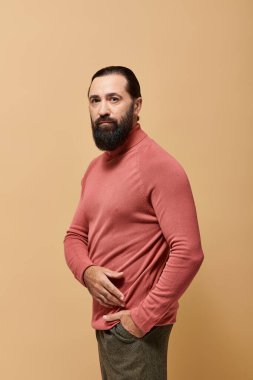Portre, odaklanmış ve sakallı yakışıklı adam bej arka planda pembe balıkçı yaka kazak giyiyor.