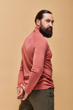 Sakallı yakışıklı ve ciddi bir adam pembe balıkçı yaka kazakla bej arka planda poz veriyor.