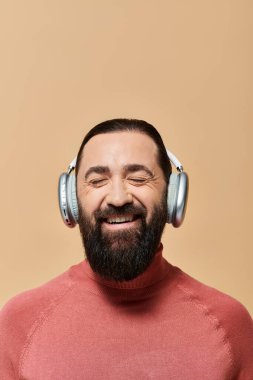 Balıkçı yakalı mutlu sakallı adam kablosuz kulaklıkla müzik dinliyor, neşeyle gülümsüyor.
