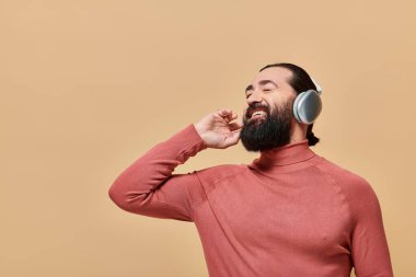 Balıkçı yaka kazaklı neşeli sakallı adam kablosuz kulaklıkla müzik dinliyor, bej renkte gülümsüyor.