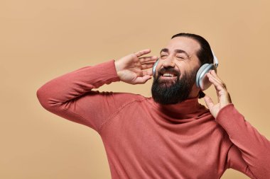Balıkçı yaka kazaklı, sakallı ve pozitif bir adam kablosuz kulaklıkla müzik dinliyor.