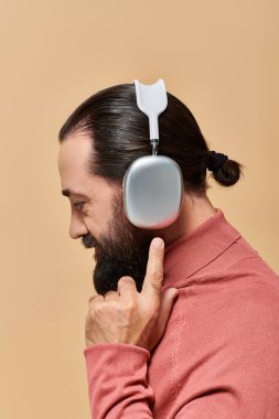 Yan görüş, balıkçı yaka kazaklı sakallı adam bej arka planda kablosuz kulaklıkla müzik dinliyor.