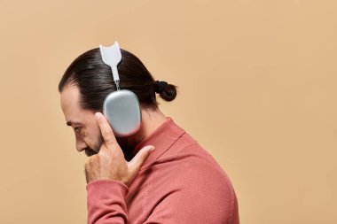 Yan görüş, balıkçı yaka kazaklı sakallı adam bej renkte kablosuz kulaklıkla müzik dinliyor.