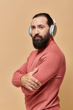 Pembe balıkçı yaka kazaklı sakallı yakışıklı adam bej kulaklıklı kablosuz kulaklıkla müzik dinliyor.