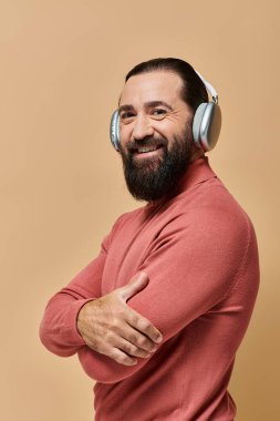 Balıkçı yaka kazaklı, kablosuz kulaklıklı, bej arka planda müzik dinleyen pozitif ve sakallı bir adam.