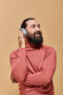 Güler yüzlü ve sakallı balıkçı yaka kazaklı adam kablosuz kulaklıkla müzik dinliyor, bej arka plan