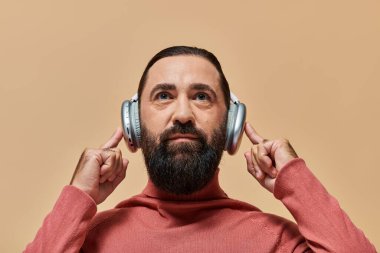 Balıkçı yaka kazaklı, sakallı ve yakışıklı adam bej renkte kablosuz kulaklıkla müzik dinliyor.