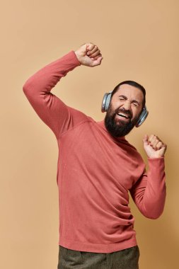 Balıkçı yaka kazaklı heyecanlı sakallı adam kablosuz kulaklıklı, bej arka planda müzik dinliyor.