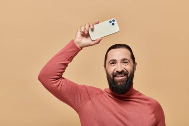 Balıkçı yaka kazaklı mutlu sakallı adamın portresi bej arka planda akıllı telefon tutuyor.