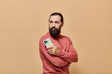 Balıkçı yaka kazaklı yakışıklı sakallı adamın portresi bej arka planda akıllı telefon tutuyor.
