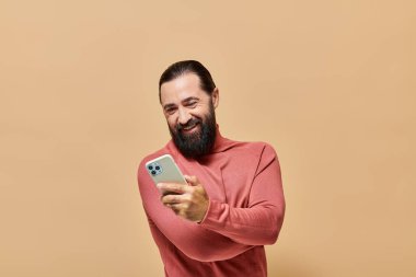 Balıkçı yaka kazaklı neşeli sakallı adamın portresi bej arka planda akıllı telefon tutuyor.
