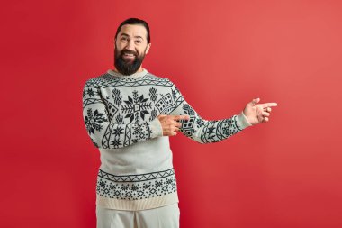 happy bearded man in Christmas sweater with ornament pointing away on red backdrop, winter holidays clipart