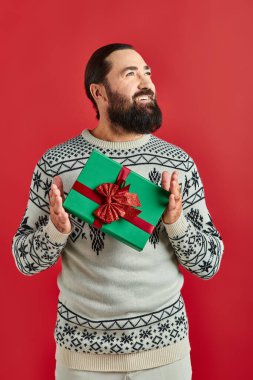 Kırmızı arka planda Noel hediyesi tutan kışın giydiği süveterli mutlu sakallı adam.
