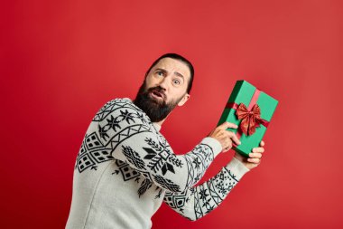 Kırmızı arka planda Noel hediyesini tutan kışın giydiği süveterli kafası karışık sakallı adam.