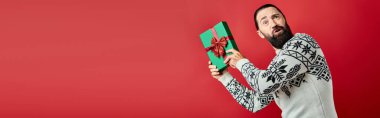 Kış süveteri giymiş kafası karışmış sakallı bir adam elinde Noel hediyesiyle kırmızı pankartta