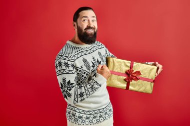 Kış süveteri içindeki neşeli sakallı adam elinde Noel hediyesiyle kırmızı arka planda.