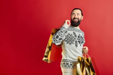 Süslü süveter giymiş sakallı, kırmızı arka planda alışveriş torbaları tutan, Noel hediyeli hayret verici bir adam.