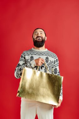 Süslü, sakallı bir adam kırmızı arka planda alışveriş torbaları tutuyor, Noel zamanı.