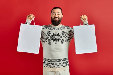 Noel süveteri giymiş neşeli sakallı adam kırmızı arka planda alışveriş torbaları tutuyor, tatil satışları