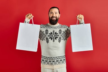 cheerful bearded man in Christmas sweater holding shopping bags on red backdrop, holiday sales clipart