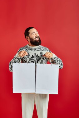 Noel süveteri giymiş pozitif sakallı bir adam kırmızı arka planda alışveriş torbaları tutuyor, tatil satışları