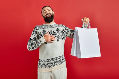 Noel süveteri giymiş pozitif sakallı bir adam kırmızı arka planda alışveriş torbaları tutuyor, bayram neşesi