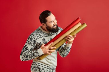 happy bearded man in winter sweater holding colorful gift paper on red backdrop, Merry Christmas clipart