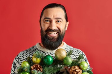 joyful bearded man in winter sweater holding decorated Christmas wreath with baubles on red backdrop clipart