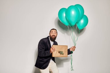 Mavi balonlar tutan, takım elbiseli mutlu sakallı adam ve gri arka planda Noel hediyesini paketleyen adam.