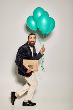 positive bearded man in formal wear holding blue balloons and Christmas  present on grey backdrop clipart