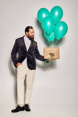 happy bearded man in suit holding blue balloons and Christmas present on grey, hand in pocket clipart