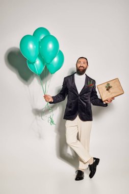Resmi kıyafetli gülümseyen sakallı adam Noel hediyesini ve gri arka planda mavi balonları tutuyor.