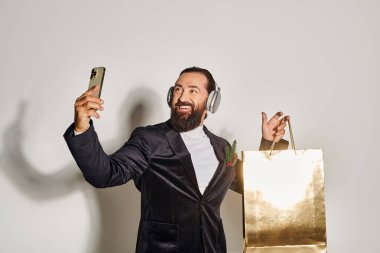 Kulaklıklı mutlu sakallı adam akıllı telefondan selfie çekiyor ve gri arka planda alışveriş çantası alıyor.