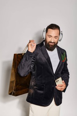 Kablosuz kulaklıklı neşeli sakallı adam gri arka planda akıllı telefon ve alışveriş çantası tutuyor.